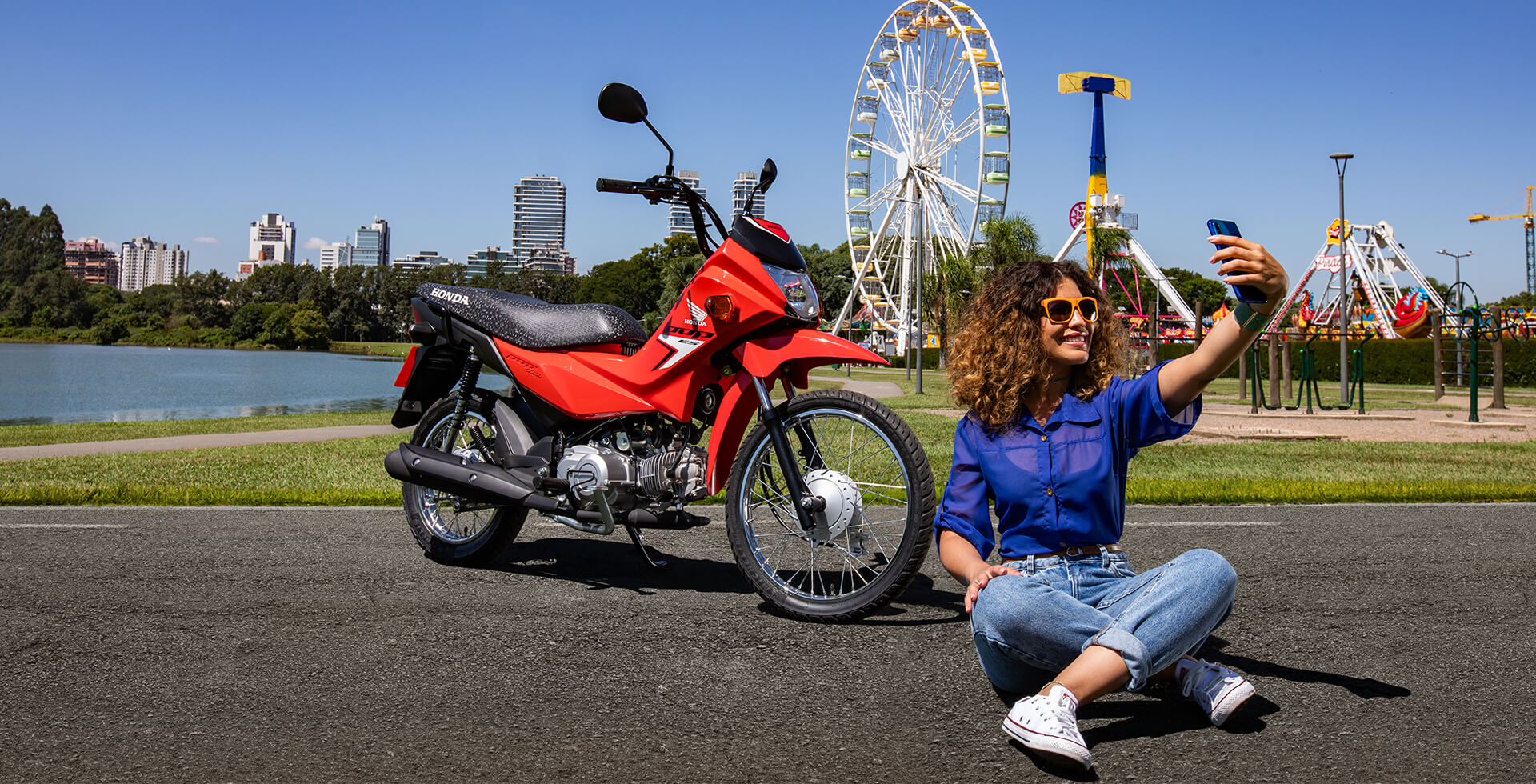 Mulher sentada ao chão em parque de diversões, tirando uma foto selfie com a motocicleta Honda Pop 110i ES Vermelho Maceio Red