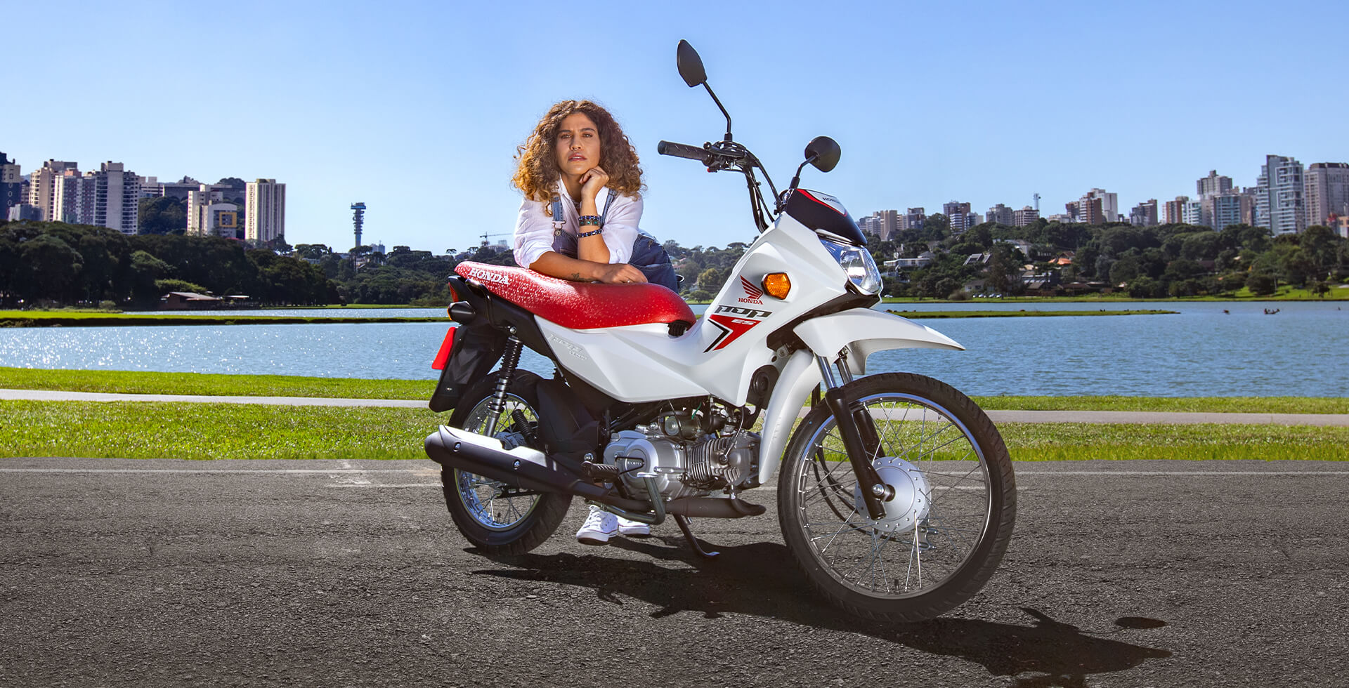 Mulher com a mão ao rosto apoiada sobre a Moto Honda Pop 110i ES na cor Branco Ross White com paisagem de lago e prédios ao fundo