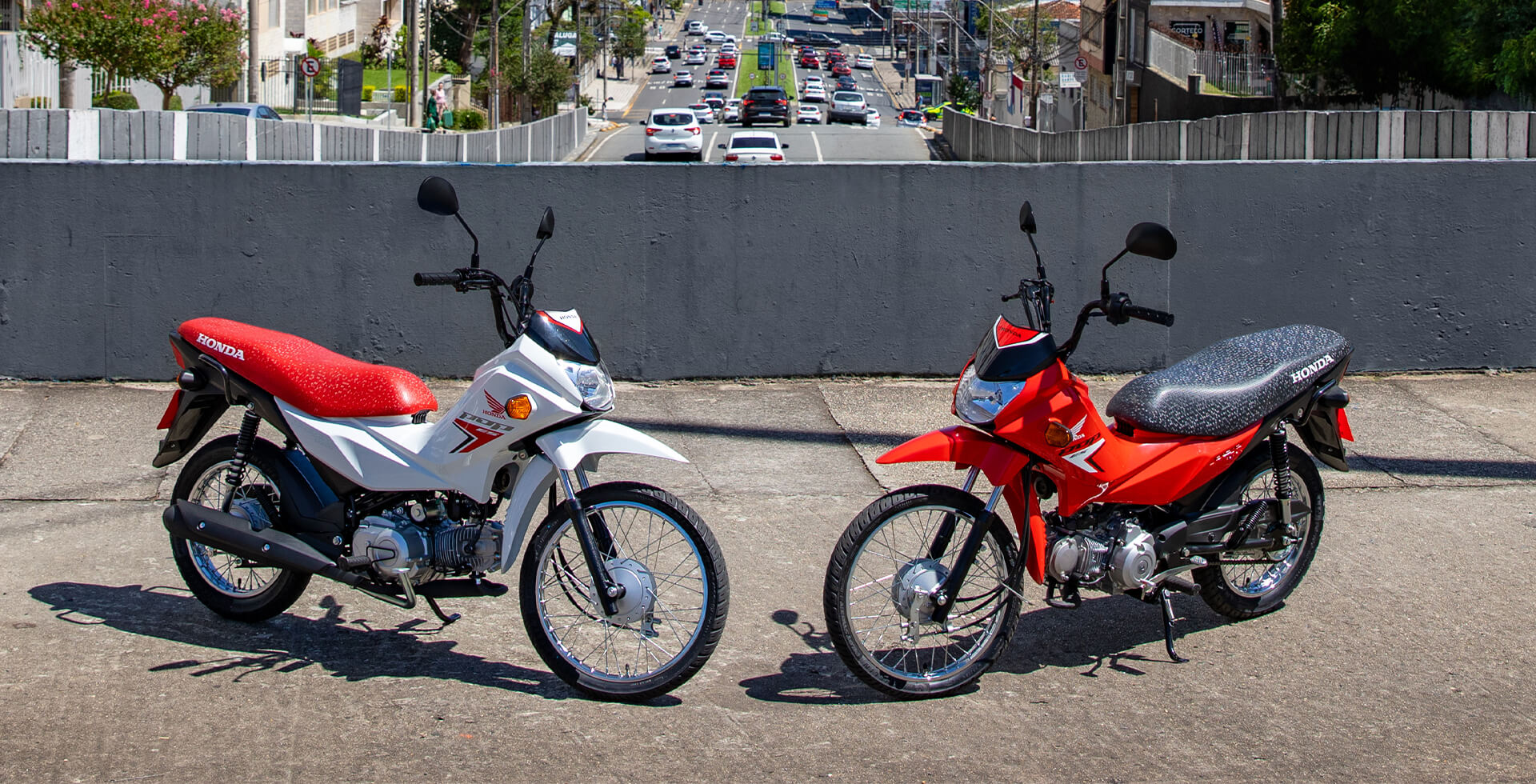 Duas Motos Pop 110i ES nas cores Branco Ross White e Vermelho Maceio Red estacionadas em cenário urbano.