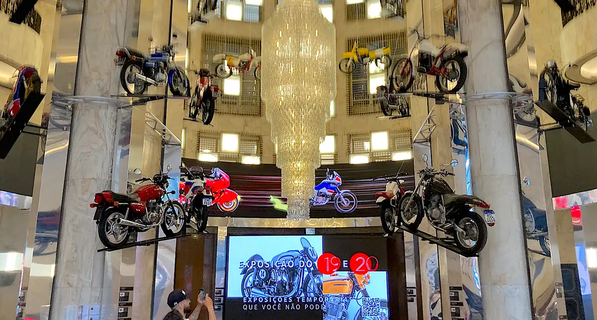 Vista do hall principal da exposição "Duas Rodas e uma Nação" com motocicletas suspensas e uma tela de informações