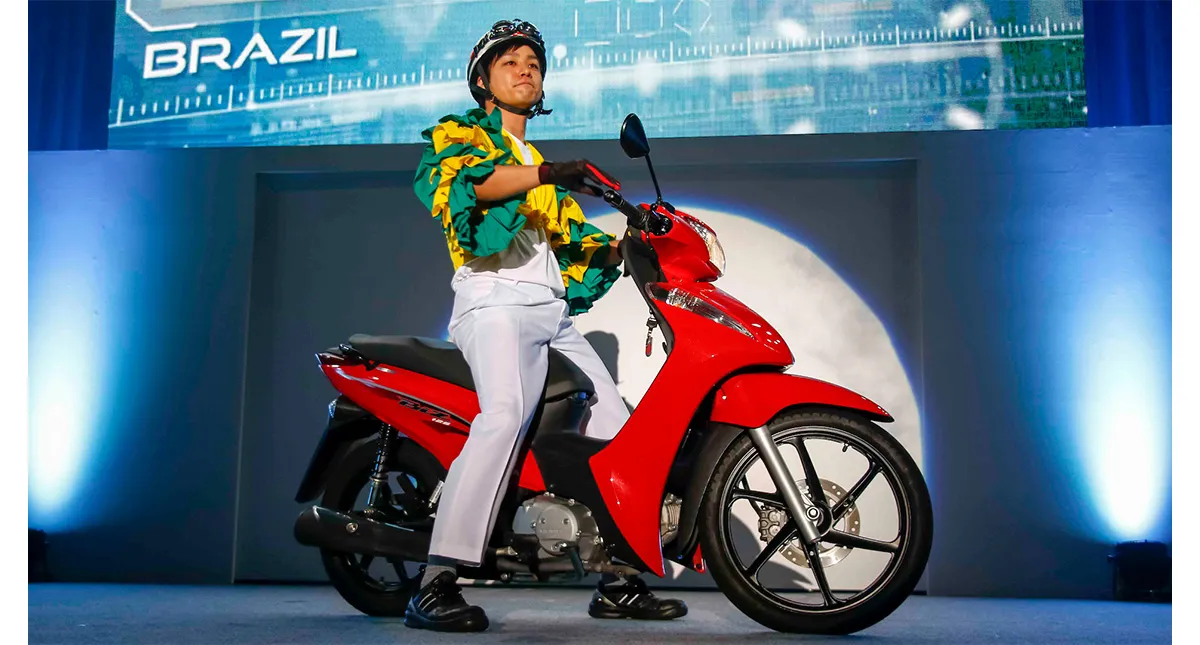 Apresentador vestido com traje colorido em cima de uma motocicleta Honda Biz vermelha durante evento de lançamento no Brasil.