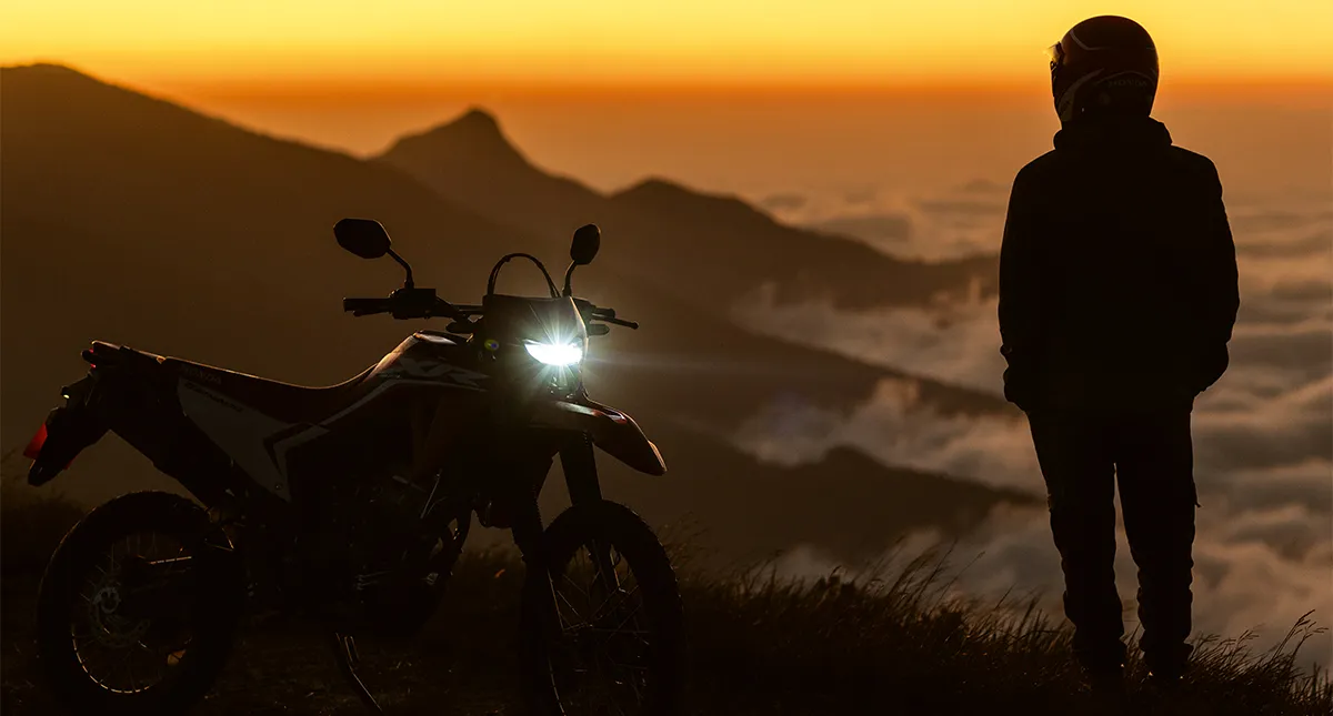 Motocicleta Honda XR 300L Tornado 2025 no anoitecer com farol aceso e homem ao lado