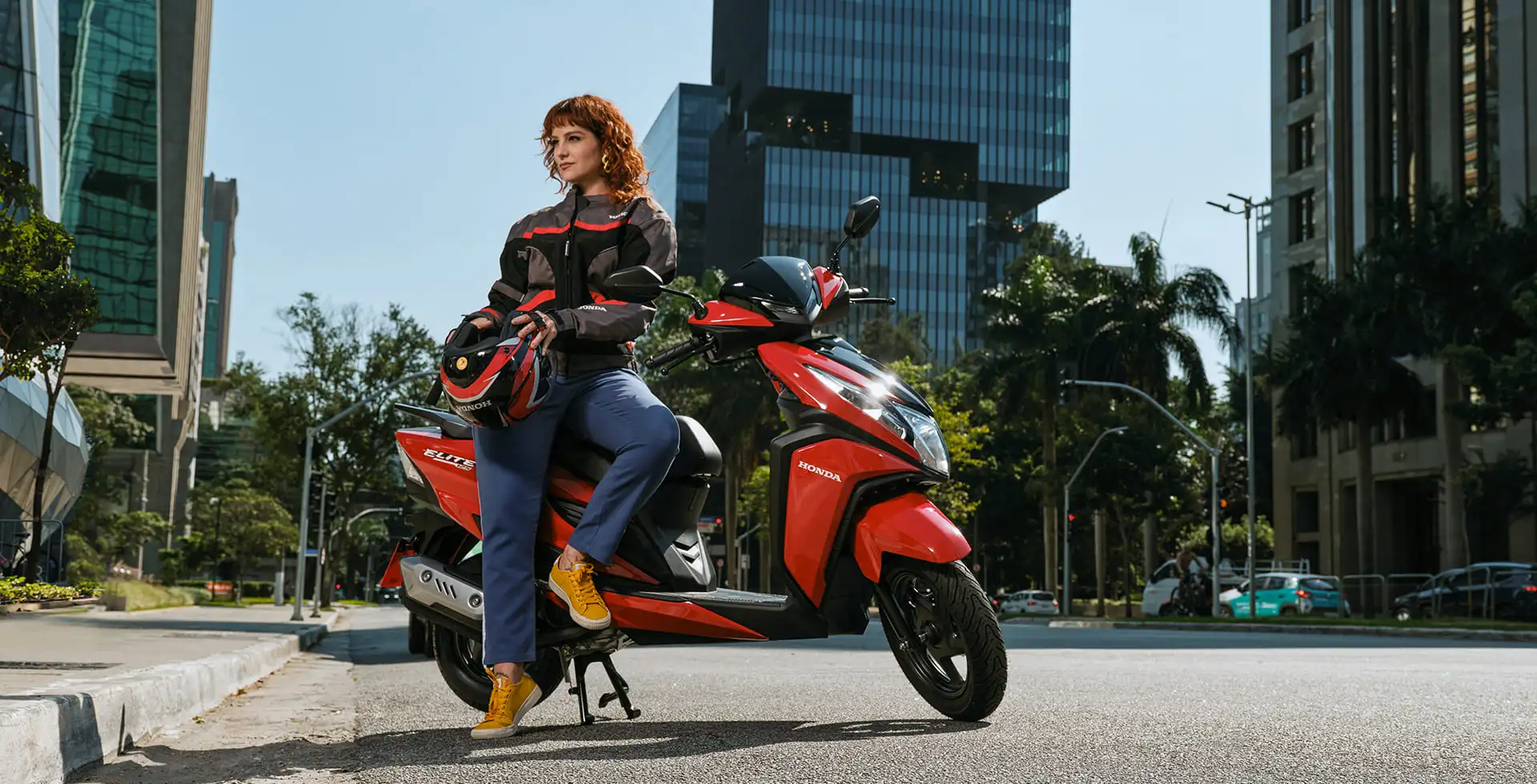 Mulher encostada ao lado da moto Honda Elite 125 Vermelho Maceio Red em cenário urbano
