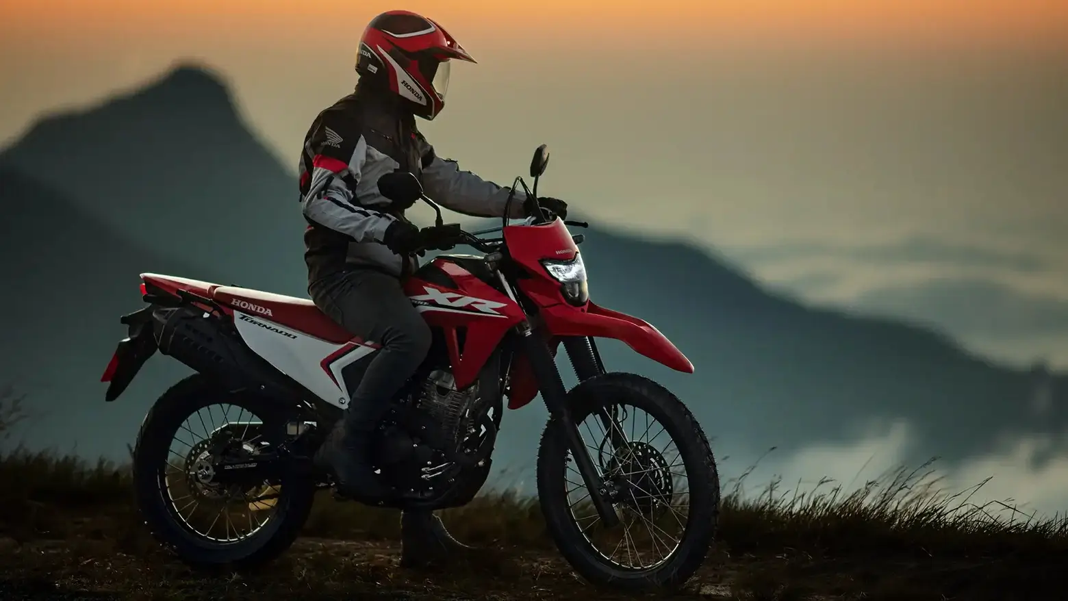 Piloto em sua Honda XR 300L Tornado pilotando em trilha com montanhas ao fundo