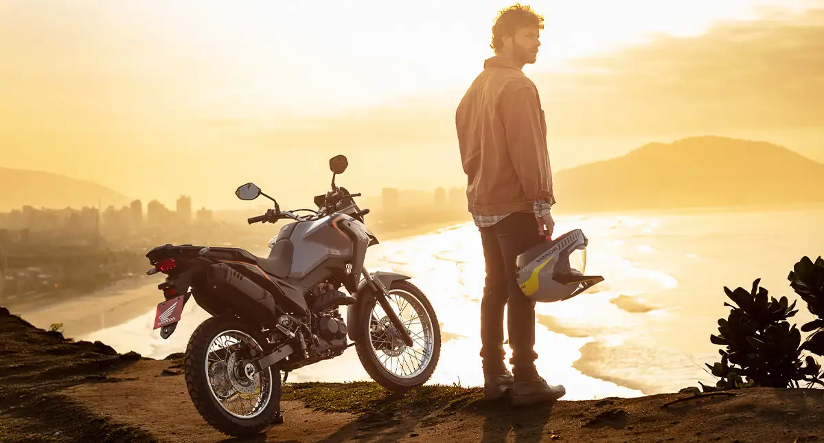 Homem com capacete na mão ao lado de motocicleta Honda XRE 190 2025 e praia ao fundo