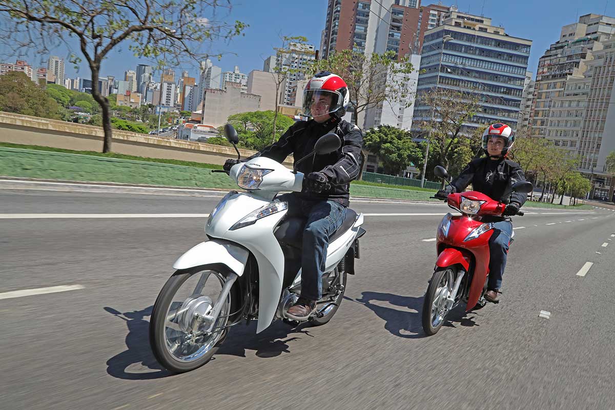 Casal andando rua de cidade com scooter Honda Biz