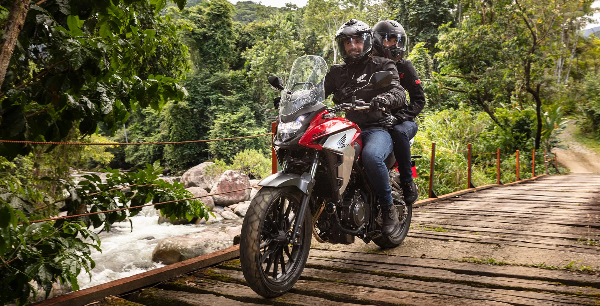 Homem com garupa em moto Honda atravessando ponte de madeira