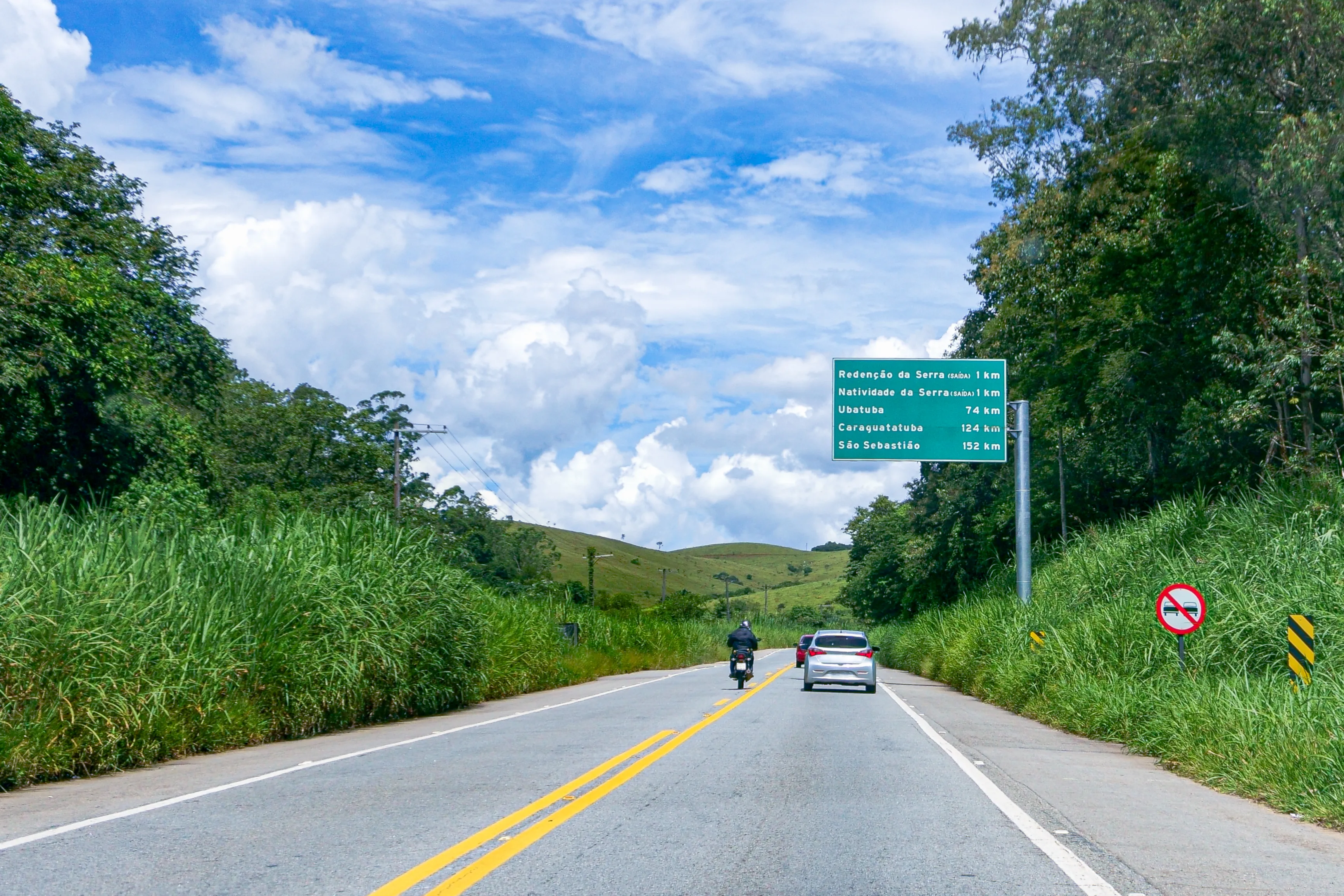 Moto e carro em estrada de asfalto com placa que sinaliza distância de cidades