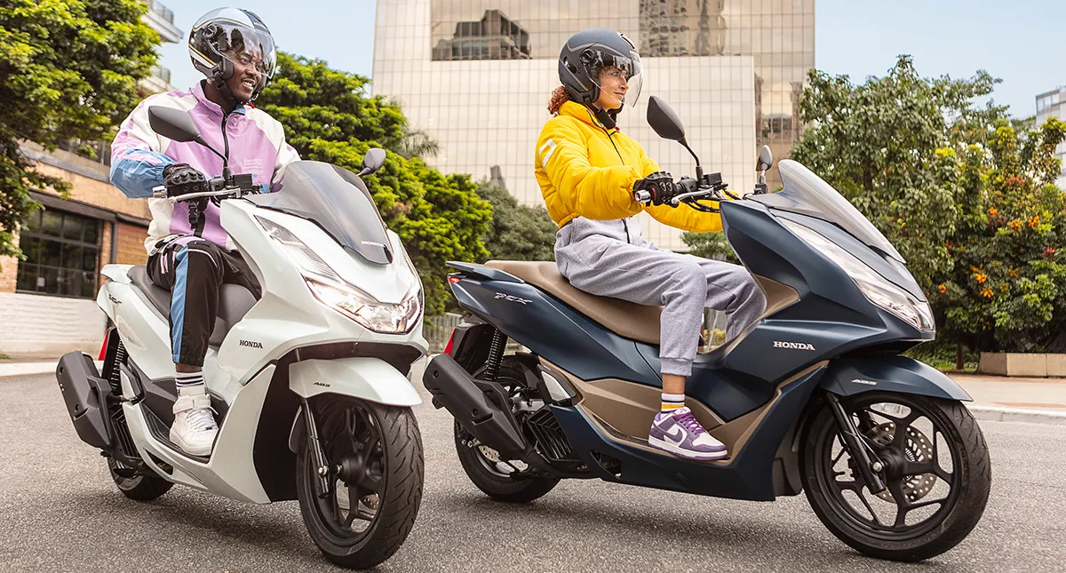 Homem negro de jaqueta roxa sentado em moto Honda PCX branca e Mulher branca de jaqueta amarela sentada em scooter PCX azul