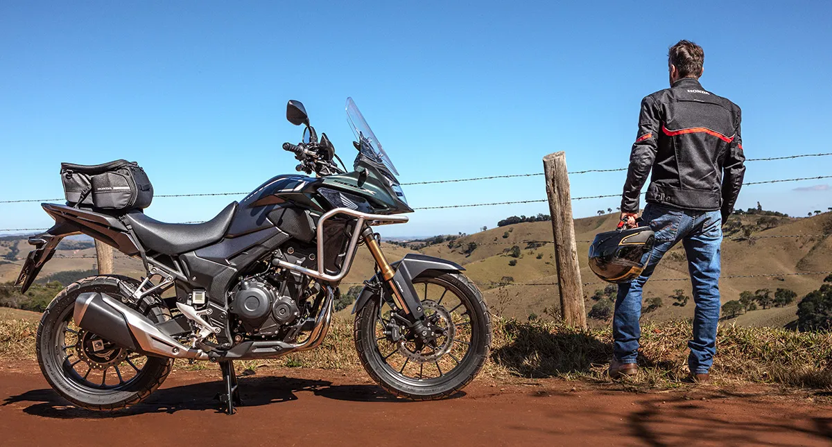 Homem Segurando Capacete na Mão Olhando Paisagem com Moto Honda CB 500X ao Lado