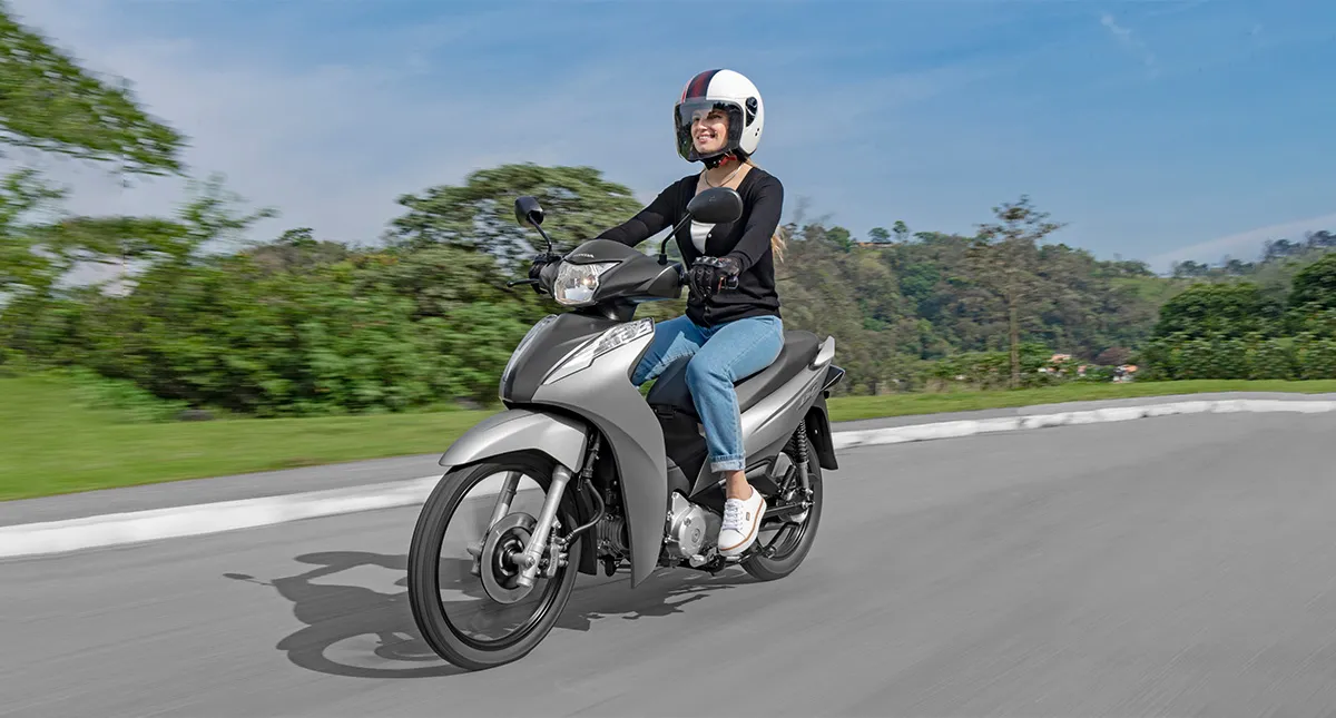 Mulher pilotando Honda Biz 125 cinza em estrada com árvores ao fundo