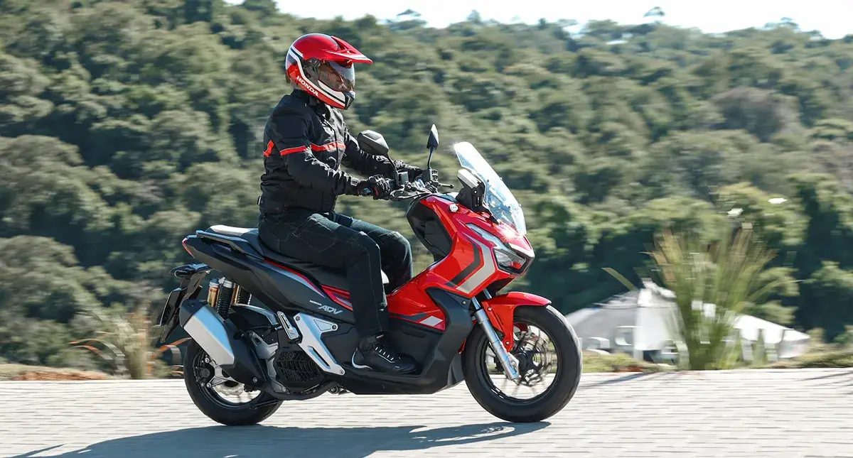 Moto Honda Scooter ADV em movimento, cor vermelha, preta e branca, vista da lateral direita, com motocilista vestindo preto e vermelho, capacete vermelho, com cenário de vegetação, estrada de ladrilhos