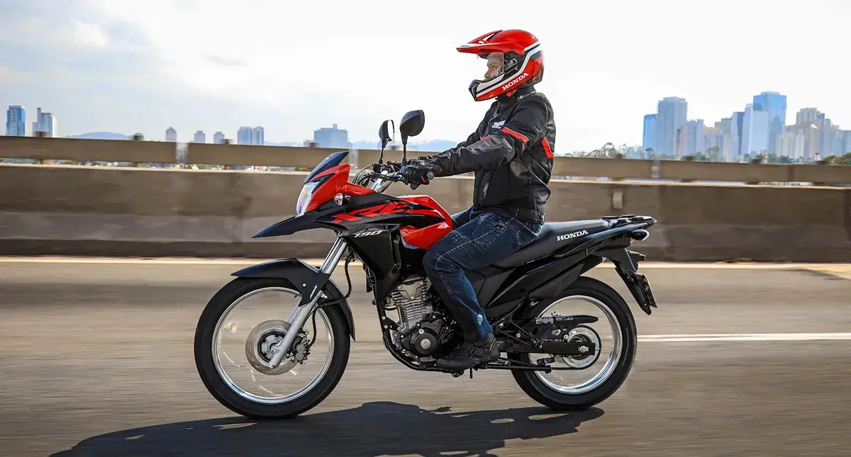 Moto Honda XRE 190 em movimento, cor vermelha e preta, vista da lateral esquerda, com motociclista vestindo preto e vermelho, capacete vermelho, em estrada asfaltada com prédios ao fundo