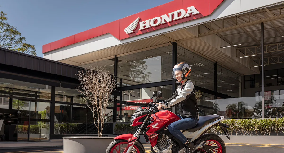Mulher saindo pilotando sua moto vermelha de uma concessionária Honda