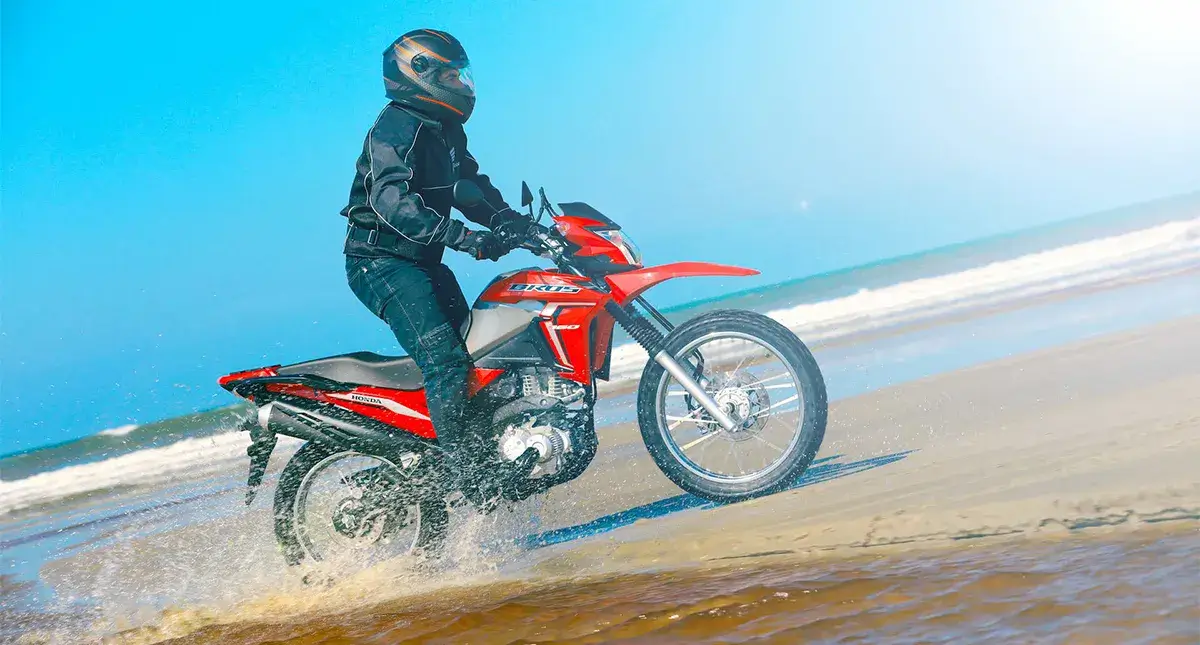 Piloto em pé e em movimento na sua Moto Honda NXR 160  Bros vermelha a beira mar 