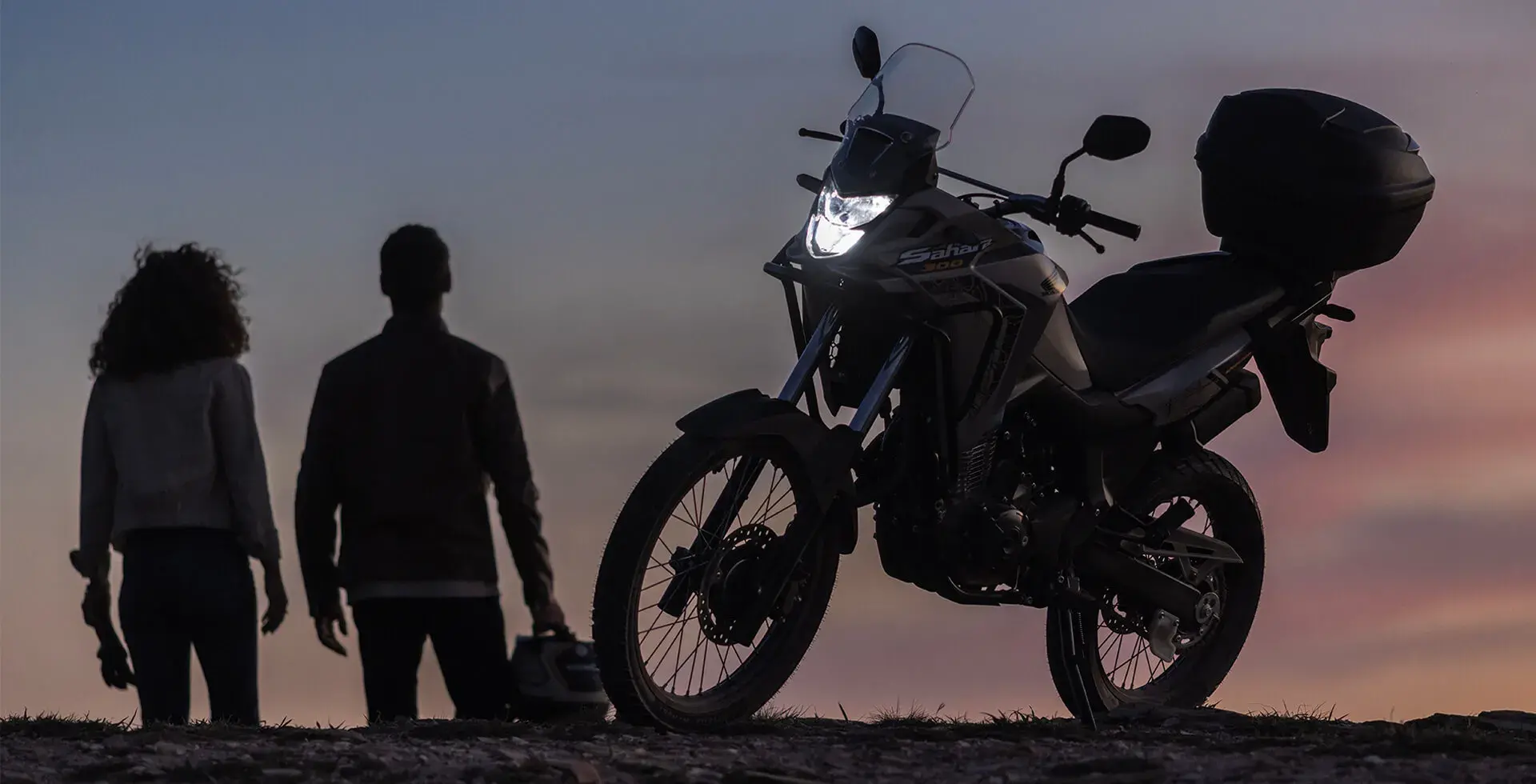 Homem e mulher lado a lado observando o pôr do sol ao lado da Honda Sahara 300