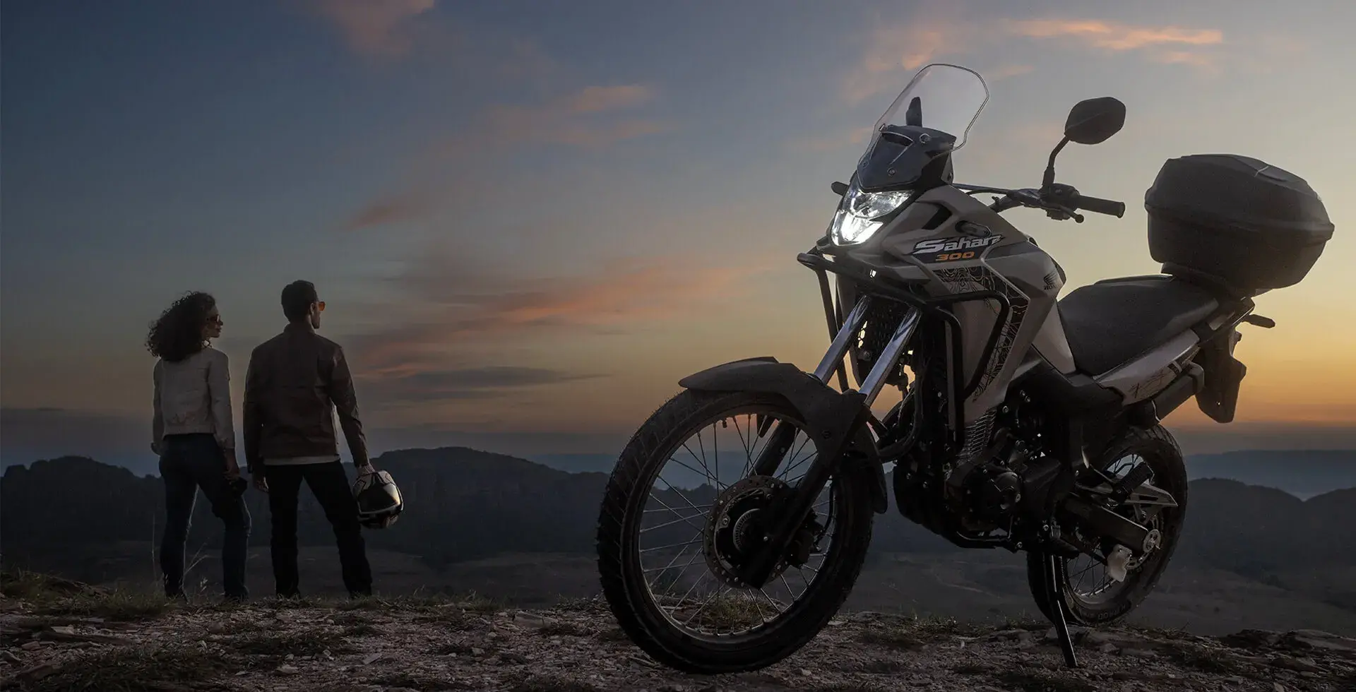 Homem e mulher lado a lado observando o pôr do sol ao lado da Honda Sahara 300