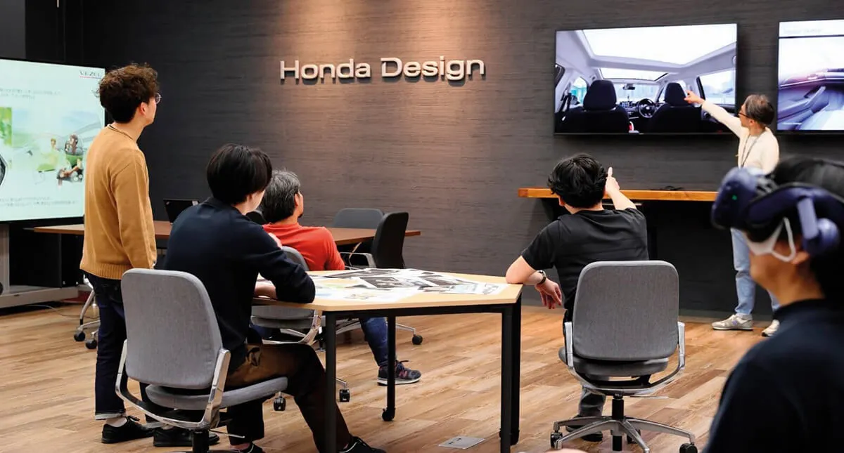 Colaboradores em sala de reunião olhando para uma tela de computador e discutindo sobre os designs da Honda, ao fundo na parede escrito Honda Design