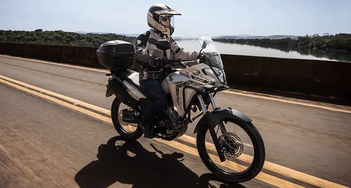 Homem em moto Honda Sahara 300 Adventure atravessando ponte de asfalto sobre rio