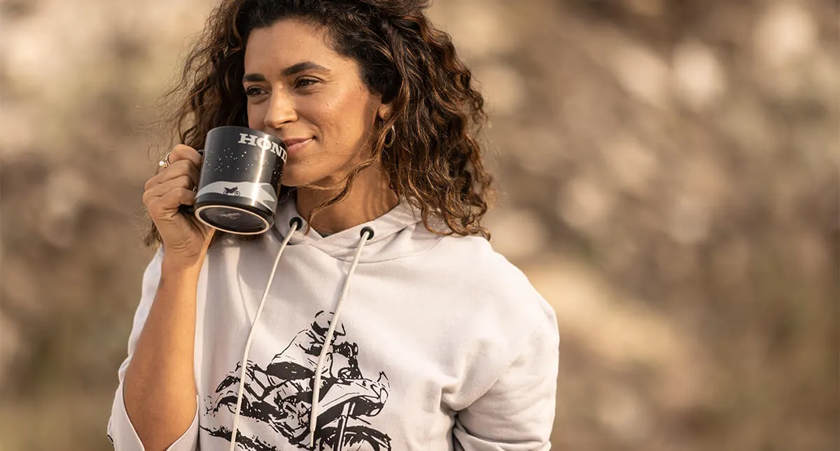 Mulher sorrindo com caneca preta de estampa honda em mãos e moletom branco com estampa de um piloto em sua moto