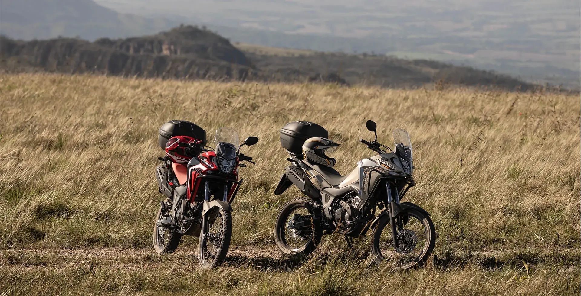 Motos Honda Sahara 300 Adventure e Rally com capacete no banco em paisagem de mato e montanha 