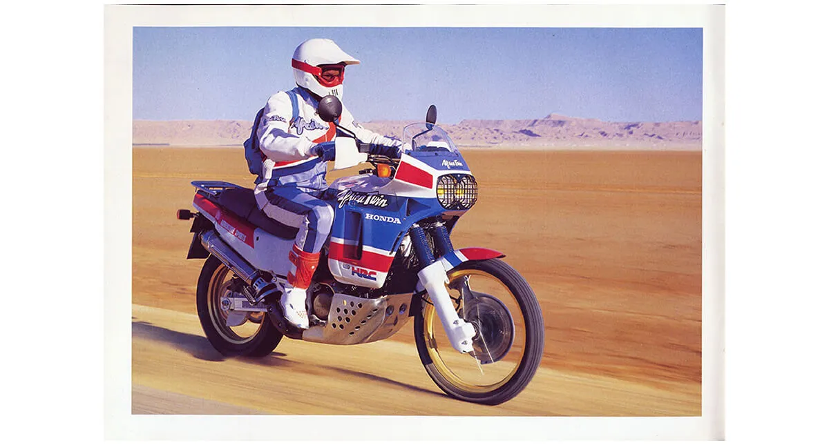 Piloto de roupa e capacete branco em motocicleta Honda XRV 650 no deserto