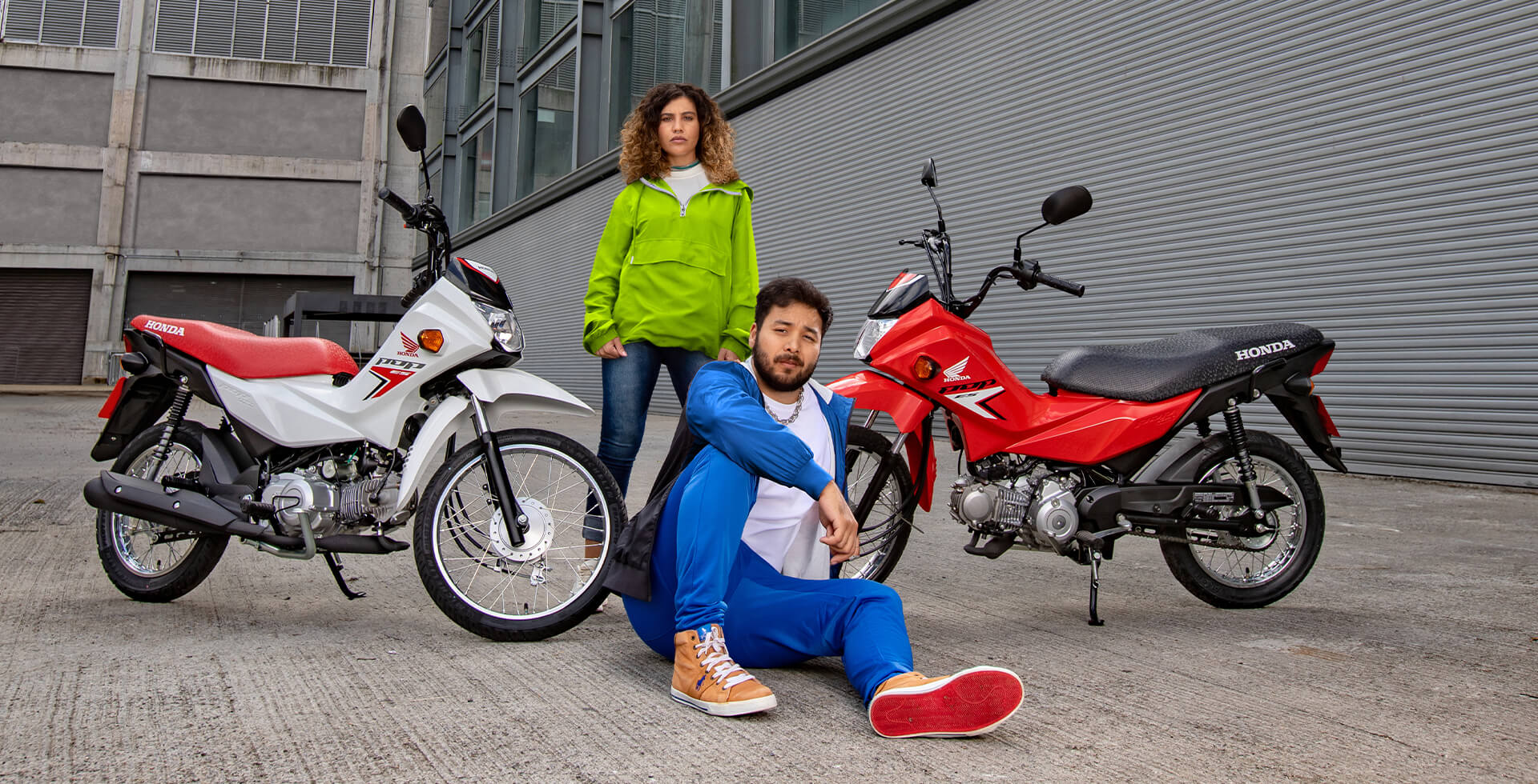 Casal em cenário urbano fazendo pose para foto e entre as motocicletas Honda Pop 1101 ES nas cores Branco Ross White e Vermelho Maceio Red