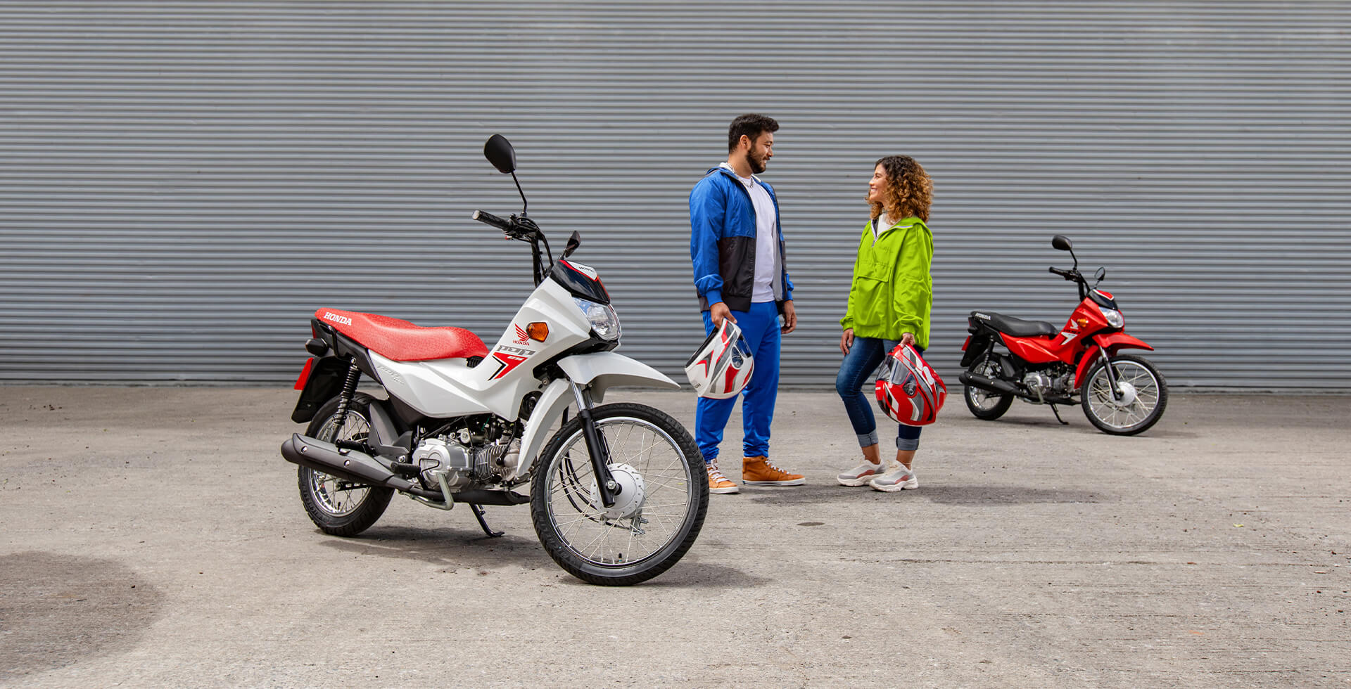 Casal em pé se olhando e segurando capacetes Honda entre duas Motocicletas Pop ES 110i nas cores Branco Ross White e Vermelho Fighting Red