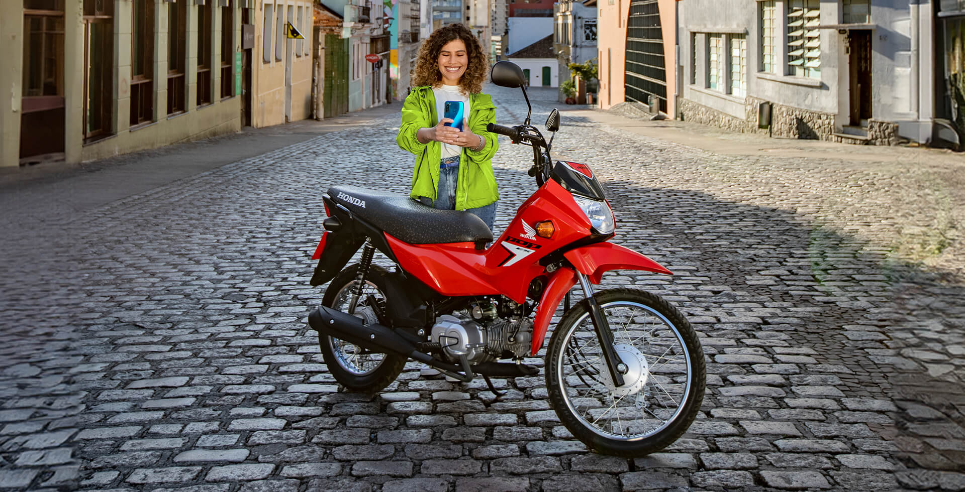 Mulher em Pé em Rua de Paralelepípedo Tirando Foto Selfie com a Moto Honda Pop 110i ES Vermelho Fighting Red
