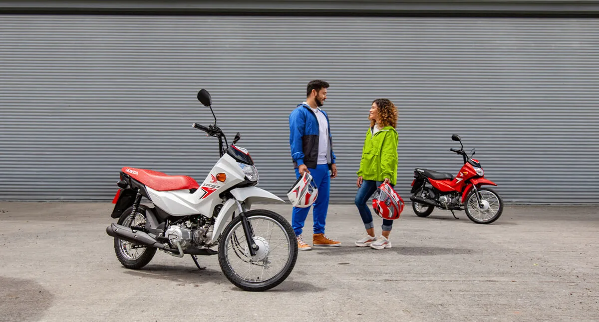 Homem e mulher se olhando entre as motocicletas Pop 110i ES Branco Ross White e Pop 110i ES Vermelho - Fighting Red Red enquanto seguram capacetes Honda