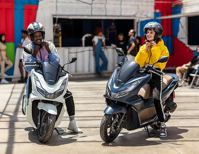 Homem em moto PCX branca e mulher em moto PCX azul abrindo fivela de capacete. 